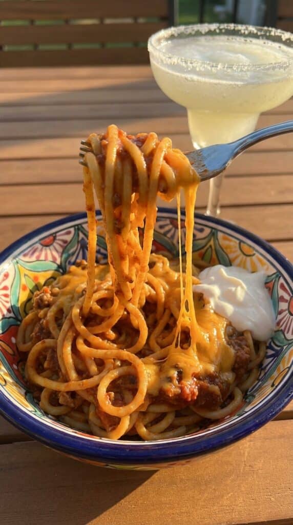 A close-up of a fork twirling cheesy taco spaghetti from a bowl, with a cheese pull and a dollop of sour cream.