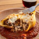 A close-up of a fork cutting into a stuffed pasta shell, revealing a ground beef and ricotta filling with a long cheese pull.