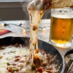 A close-up of a tortilla chip scooping into a hot layered bean and cheese dip, creating a massive, stringy cheese pull.