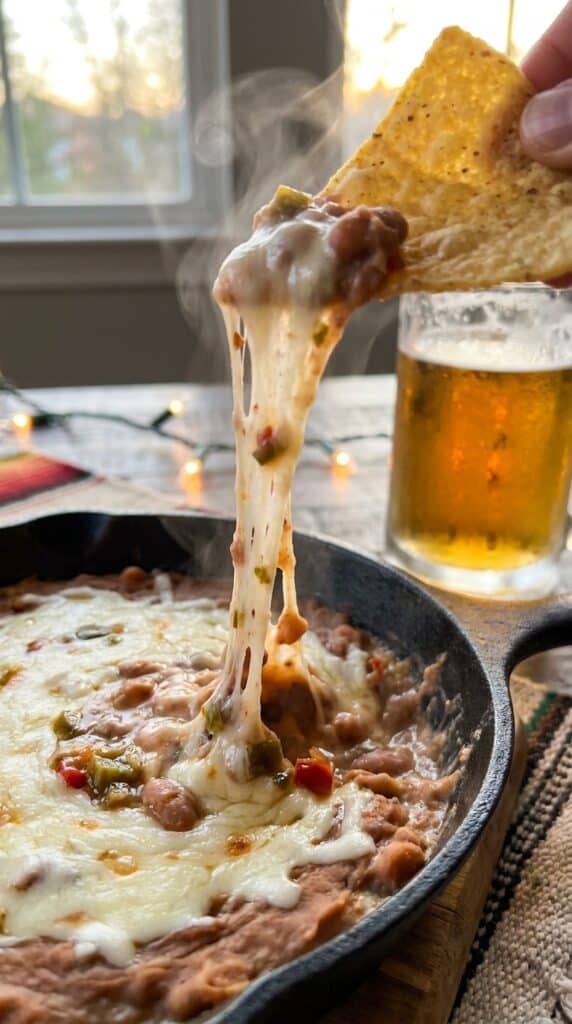 A close-up of a tortilla chip scooping into a hot layered bean and cheese dip, creating a massive, stringy cheese pull.