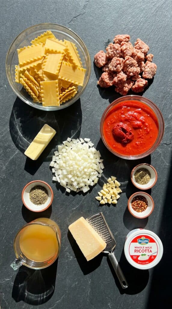 An overhead flat lay showing clusters of raw ingredients for lasagna soup: broken lasagna noodles, crumbled Italian sausage, diced onion, tomato paste, canned tomatoes, broth, spices, and a wedge of Parmesan cheese on a slate board.