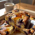 A close-up of a spatula lifting a slice of blueberry French toast casserole while maple syrup is being drizzled on top, revealing a custardy interior.