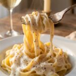 A close-up of a silver fork twirling wide fettuccine noodles coated in a thick, dripping white cream sauce, with a glass of wine in the background.