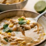 A close-up of a soup spoon holding shredded chicken, white beans, and corn coated in a creamy broth, with tortilla chips in the background.