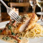 A close-up of a fork cutting into golden chicken topped with creamy sun-dried tomato and spinach sauce, served over angel hair pasta.