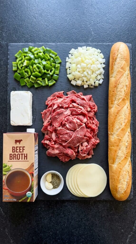 A flat lay showing shaved beef, green bell peppers, onions, beef broth, cream cheese, provolone, and a baguette on a dark slate board.
