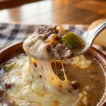A close-up of a soup spoon breaking through melted cheese to lift a bite of shaved beef and peppers from a bowl of creamy soup.