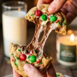 Two hands pulling apart a thick holiday chocolate chip cookie, showing a gooey melted chocolate center and red and green candies.