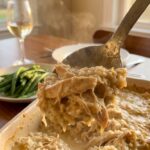 A close-up of a serving spoon lifting a scoop of hot, creamy chicken and rice casserole with steam rising.