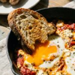 A close-up of a piece of crusty bread dipping into a runny egg yolk and melted feta cheese in a skillet, with chicken sausage in the background.