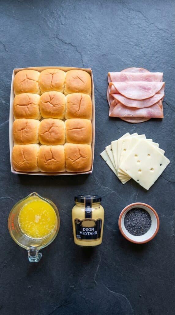 A flat lay showing a pack of sweet rolls, sliced ham, sliced Swiss cheese, melted butter, Dijon mustard, and poppy seeds on a dark slate board.