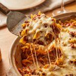 A close-up of a spatula lifting a hearty serving of layered beef, noodle, and cheese casserole with a dramatic cheese pull.