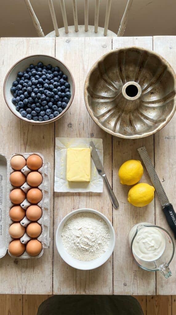 A flat lay showing fresh blueberries, a metallic bundt pan, butter, lemons, eggs, flour, and sour cream on a wooden board.
