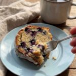 A close-up of a fork cutting into a thick slice of blueberry bundt cake on a blue plate, revealing berries inside, with coffee in the background.