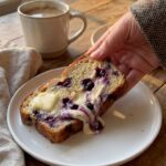A close-up of a hand holding a thick slice of blueberry cream cheese bread with melting butter, with a coffee mug in the background.