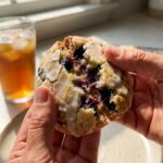 A close-up of hands breaking a glazed blueberry lemon cookie in half, revealing the soft interior and jammy baked blueberries.
