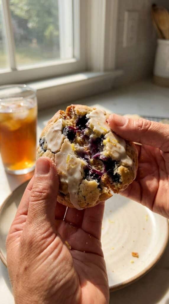 A close-up of hands breaking a glazed blueberry lemon cookie in half, revealing the soft interior and jammy baked blueberries.
