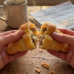 A close-up of hands pulling apart a warm sausage crescent roll, revealing a creamy, cheesy sausage filling inside, with coffee in the background.