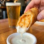 A close-up of a hand dipping a crispy buffalo chicken egg roll, showing the cheesy interior, into a bowl of ranch dressing.
