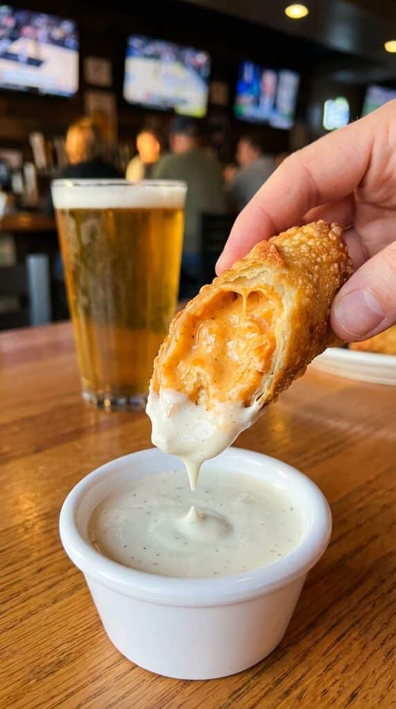 A close-up of a hand dipping a crispy buffalo chicken egg roll, showing the cheesy interior, into a bowl of ranch dressing.