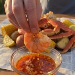 A close-up of a hand dipping a piece of shrimp into a small bowl of spicy melted garlic butter, with a seafood boil in the background.