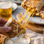 A close-up of a hand dipping a crispy, cheesy beef wrap into a bowl of sour cream, with a long cheese pull.