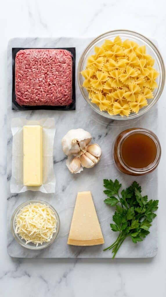 A flat lay showing raw ground beef, dry bowtie pasta, butter, garlic cloves, broth, mozzarella, and parsley on a marble board.