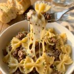 A close-up of a fork lifting a bite of bowtie pasta and beef with a long string of melted cheese stretching from a bowl.