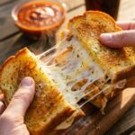 A close-up of hands pulling apart a hot garlic bread chicken sandwich, creating a massive stretch of melted cheese.