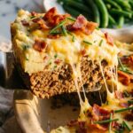 A close-up of a spatula lifting a square slice of meatloaf and potato casserole, showing the distinct beef and potato layers with a cheese pull.