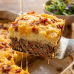 A close-up of a spatula lifting a slice of meatloaf casserole, showing distinct layers of meat, mashed potatoes, and melted cheese.