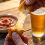 A close-up of a golden fried mozzarella bite being pulled apart by two hands, revealing a massive, stretchy cheese pull.