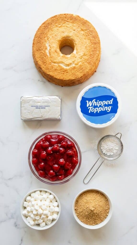 A flat lay showing a whole angel food cake, cream cheese, whipped topping, powdered sugar, and a bowl of cherry pie filling on a marble board.