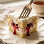 A close-up of a plated square of angel food cake and cream dessert topped with cherries, with a fork taking a bite.