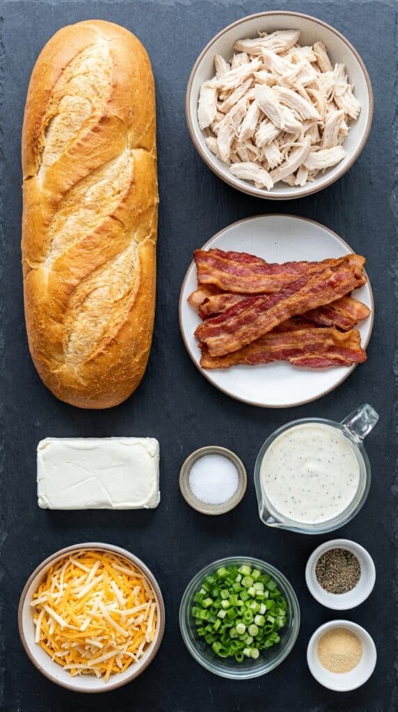 A flat lay showing a whole loaf of French bread, shredded chicken, bacon, cream cheese, ranch dressing, and shredded cheese on a dark board.