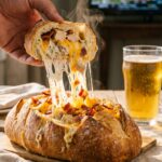 A close-up of a hand pulling a slice of stuffed bread from the loaf, creating a long, gooey cheese pull.