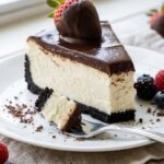 A close-up of a slice of cheesecake showing the Oreo crust, vanilla filling, chocolate ganache, and dipped strawberry, with a fork taking a bite.