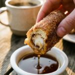 A close-up of a hand dunking a half-eaten cinnamon sugar French toast roll-up into a small bowl of maple syrup.