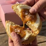 A close-up of hands breaking a giant frosted caramel cookie in half, showing the soft, dense center and dripping caramel.