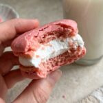 A close-up of a hand holding a bitten pink heart cake, showing the pink cake interior and thick white marshmallow filling.
