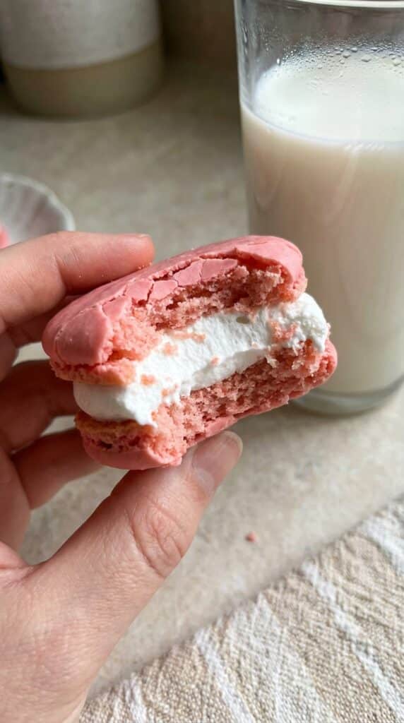 A close-up of a hand holding a bitten pink heart cake, showing the pink cake interior and thick white marshmallow filling.