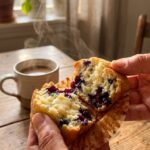 A close-up of hands pulling a warm blueberry muffin in half, showing a fluffy interior, steaming blueberries, and melting butter.