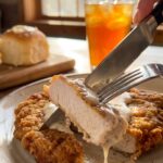 A close-up of a knife and fork cutting into a breaded pork chop smothered in peppery white gravy with bacon.