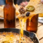 A close-up of a tortilla chip pulling gooey melted cheese loaded with beef, beans, and a jalapeño from a skillet, with frosty beers in the background.