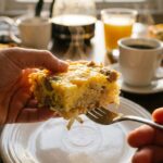 A close-up of a fork cutting into a square slice of baked breakfast casserole on a plate, showing the fluffy internal layers of egg, cheese, hash browns, and sausage.