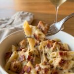 A close-up of a fork lifting a bite of creamy chicken and bacon penne pasta with a slight cheese pull from a white bowl.