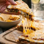 A close-up of a hand lifting a slice of Mexican pizza, showing the crispy layers and a long cheese pull.