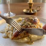 A close-up of a fork and knife cutting into pan-seared chicken covered in cream cheese sauce, served over mashed potatoes.