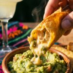 A close-up of a hand dipping a broken, crispy chicken taquito into a bowl of chunky guacamole, showing the creamy filling.