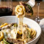 A close-up of a fork lifting a piece of tortellini, steak, and green pepper from a bowl, creating a long, gooey cheese pull.